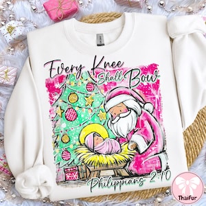 May include: White sweatshirt with a Christmas-themed graphic. The design features a Christmas tree, Santa Claus, and a baby in a manger. Text reads "Every Knee Shall Bow" and "Philippians 2:10". The graphic is in pink, green, and gold.