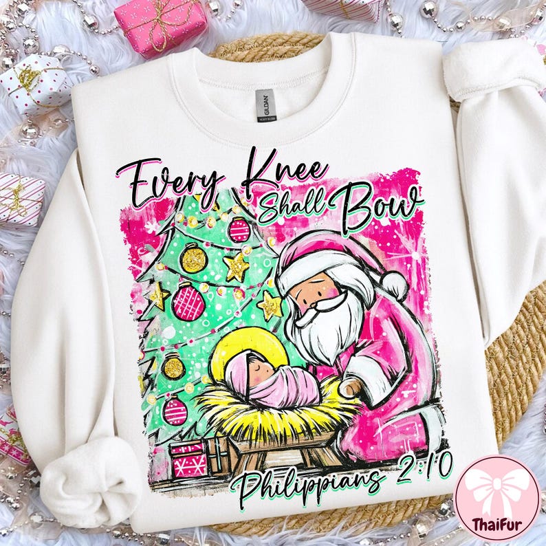 May include: White sweatshirt with a Christmas-themed graphic. The design features a Christmas tree, Santa Claus, and a baby in a manger. Text reads "Every Knee Shall Bow" and "Philippians 2:10". The graphic is in pink, green, and gold.