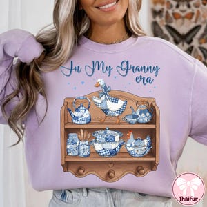 K&ouml;nnte beinhalten: Ein helllila Sweatshirt mit dem Text "In My Granny era" in blauer Schrift. Das Design zeigt ein Holzregal mit blau-wei&szlig; kariertem K&uuml;chengeschirr und einer Gans. Das Regal hat einen herzf&ouml;rmigen Ausschnitt.