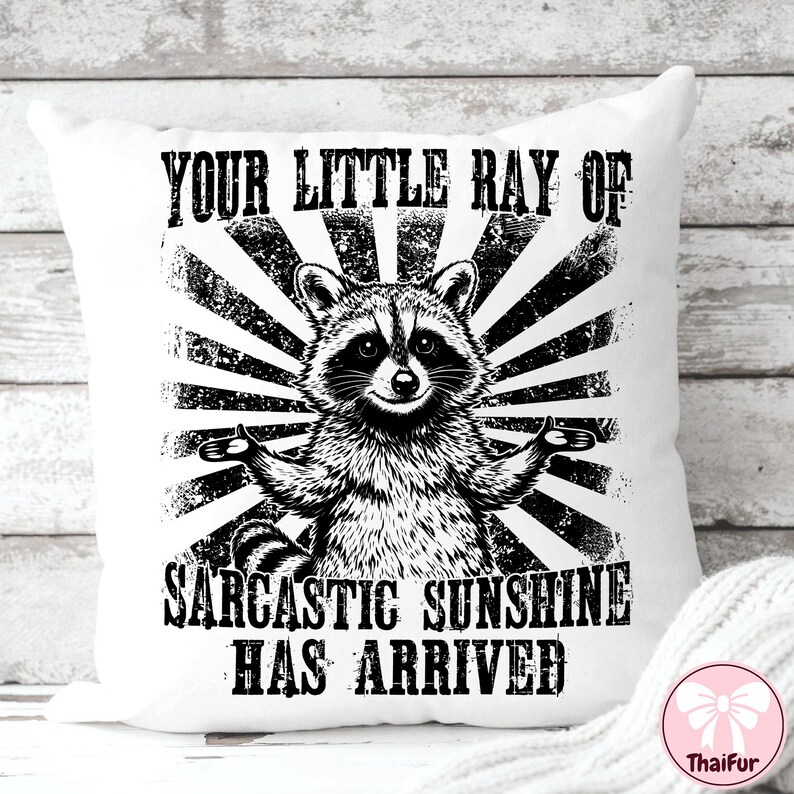 May include: White throw pillow with a black and white graphic of a raccoon with outstretched arms, set against a sunburst design. The text reads "YOUR LITTLE RAY OF SARCASTIC SUNSHINE HAS ARRIVED."