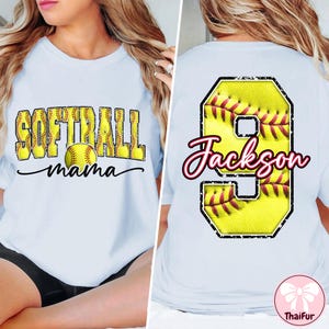 Op de afbeelding: Lichtblauw T-shirt met de tekst "SOFTBALL mama" in geel en zwart op de voorkant. De achterkant toont een groot geel honkbalnummer 9 met de naam "Jackson" in rood en zwart script. Een shirt met softbalthema.