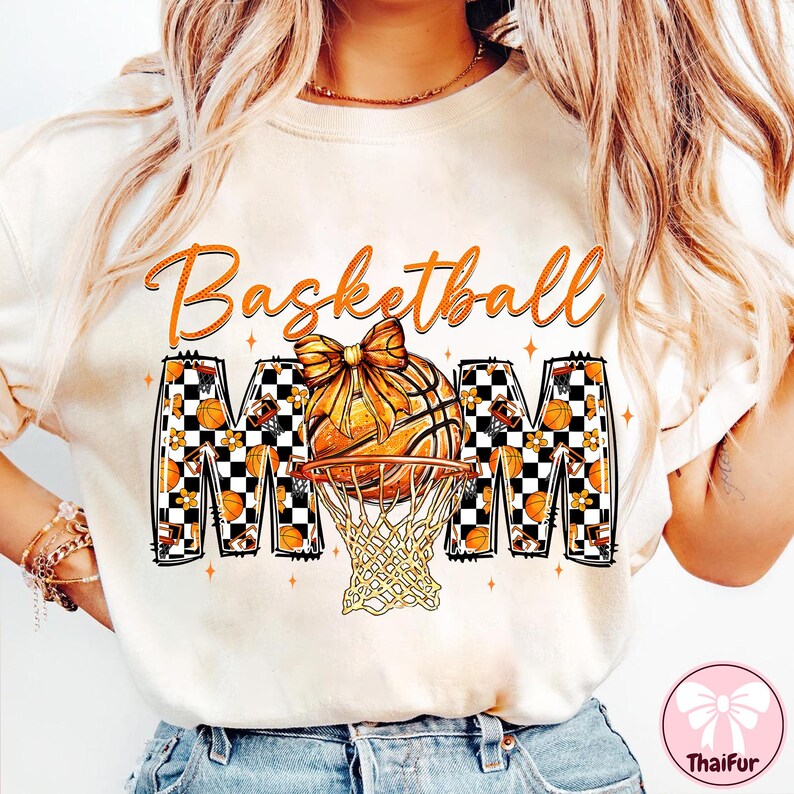 May include: Cream-colored t-shirt with the words "Basketball Mom" in orange script. The design features a basketball with a bow, a net, and the letters "MOM" in a checkered pattern with basketballs and flowers.
