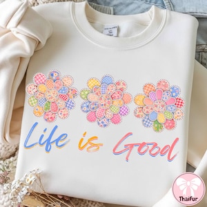 May include: Cream-colored sweatshirt featuring three floral designs made of colorful patchwork. The phrase "Life is Good" is written in blue, orange, and pink script across the front. A casual, comfortable top.