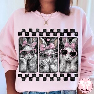 May include: Pink sweatshirt featuring a graphic of three bunnies wearing heart-shaped sunglasses and pink bows. The bunnies are in black and white, set against a black background with a checkered border.