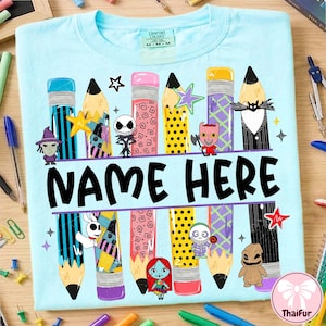 May include: A light blue t-shirt featuring a graphic of colorful pencils and characters from The Nightmare Before Christmas. The design includes the text NAME HERE in black letters, surrounded by various pencil patterns and small stars.