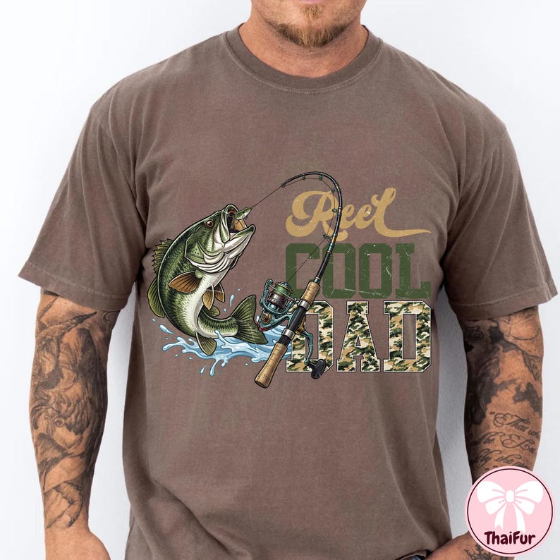 May include: Brown t-shirt with a graphic of a leaping fish, fishing rod, and the text "Reel Cool Dad" in green and camouflage. The fish is green and white, with water splashes around it.