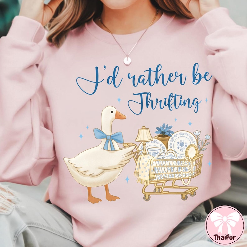 May include: Pink sweatshirt with the text "I'd rather be Thrifting". The graphic features a cartoon goose with a blue bow, pushing a shopping cart filled with vintage dishes, a lamp, and a plant. The design includes blue stars.