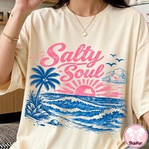 May include: Cream-colored t-shirt with a beach-themed graphic. The design features the words "Salty Soul" in pink, a blue palm tree, and a pink and orange sunset over blue waves. Three birds and clouds are also included.