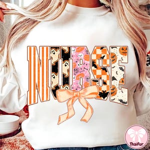 May include: White sweatshirt with the word "NURSE" in a Halloween-themed design. The letters feature orange stripes, ghosts, pink skulls, orange checkerboard, bats, and jack-o'-lanterns. A peach bow is at the bottom.