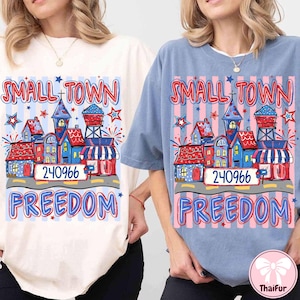 Könnte beinhalten: Zwei T-Shirts, eines in Creme und eines in Blau, zeigen eine farbenfrohe Grafik. Die Grafik zeigt eine Kleinstadt mit Häusern, einem Wasserturm und den Worten "SMALL TOWN FREEDOM" und der Nummer "240966". Sterne und Feuerwerk ergänzen das Design.