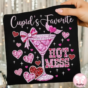 Może przedstawiać: Czarny t-shirt z graficznym wzorem przedstawiającym różowy kieliszek do koktajli, serca i napis "Cupid's Favorite HOT MESS". Projekt zawiera kokardę, skrzydła i błyszczące akcenty, idealne na Walentynki.