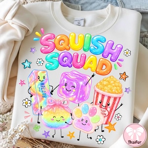 May include: A cream-colored crewneck sweatshirt featuring the colorful text SQUISH SQUAD above cartoon illustrations of a butter stick, a purple cube, a popcorn bucket, a rainbow-colored round object with a bow, and a paw print.