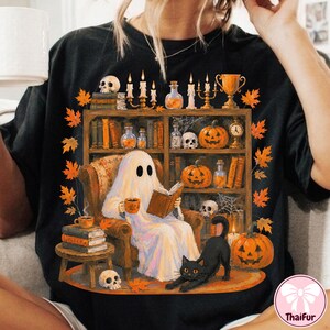 May include: A black t-shirt featuring a graphic illustration of a white ghost sitting in an armchair reading a book. The scene includes a bookshelf filled with potions, skulls, pumpkins, candles, and a black cat stretching on the floor.