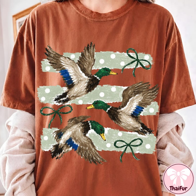 Pu&ograve; includere: Una t-shirt color ruggine con una stampa grafica di tre germani reali in volo. Il design presenta strisce orizzontali verde chiaro effetto pennellata con pois bianchi e fiocchi verde scuro posizionati tra le figure dei volatili.