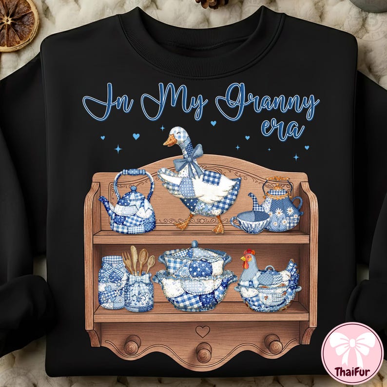 K&ouml;nnte beinhalten: Schwarzes Sweatshirt mit einem skurrilen Design mit einer Gans, Teekannen und einem Huhn, alle mit blau-wei&szlig;em Vichy-Muster verziert. Der Schriftzug "In My Granny era" steht in blauer Schrift &uuml;ber dem Regal. Ein Holzregal zeigt die Gegenst&auml;nde.