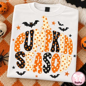 Könnte beinhalten: Ein weißes T-Shirt mit Rundhalsausschnitt und dem Schriftzug PUMPKIN SEASON in großen, abgenutzten Buchstaben mit Punktmuster in Orange, Schwarz und Creme. Das Design enthält kleine schwarze Fledermaussilhouetten und orangefarbene Sterne.