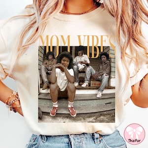 May include: White t-shirt with a graphic print of four people sitting on a porch. The text "MOM VIBES" is printed in gold above the graphic.