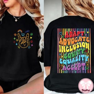 Könnte beinhalten: Schwarzes T-Shirt mit Rundhalsausschnitt und einem farbenfrohen Peace-Zeichen-Motiv auf der Vorderseite und den Worten "Adapt, Advocate, Inclusion, Equity, Equality, Accept" auf der Rückseite. Das Design umfasst Regenbogen, Herzen und Blumen.