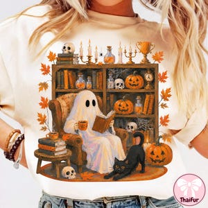 May include: A cream-colored t-shirt featuring a graphic of a ghost sitting in an armchair reading a book, surrounded by a bookshelf filled with pumpkins, skulls, candles, and potions, with a black cat and autumn leaves in the foreground.