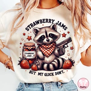 以下が含まれることがあります： 銃とイチゴジャムの瓶を持った漫画のラクーンが描かれた白いTシャツ。シャツのテキストは「Strawberry Jams But My Glock Don't.」です。