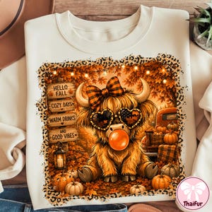 May include: A cream-colored sweatshirt featuring a graphic of a fluffy highland cow wearing a plaid bow and heart-shaped sunglasses while blowing an orange bubble. The design includes autumn leaves, pumpkins, and signs reading HELLO FALL.