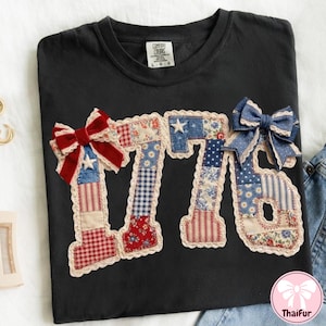 May include: Black t-shirt with a patriotic "1776" design. The numbers are made of patchwork fabric in red, white, and blue patterns, with lace trim and bows. The shirt is a Comfort Colors brand, size L-G.
