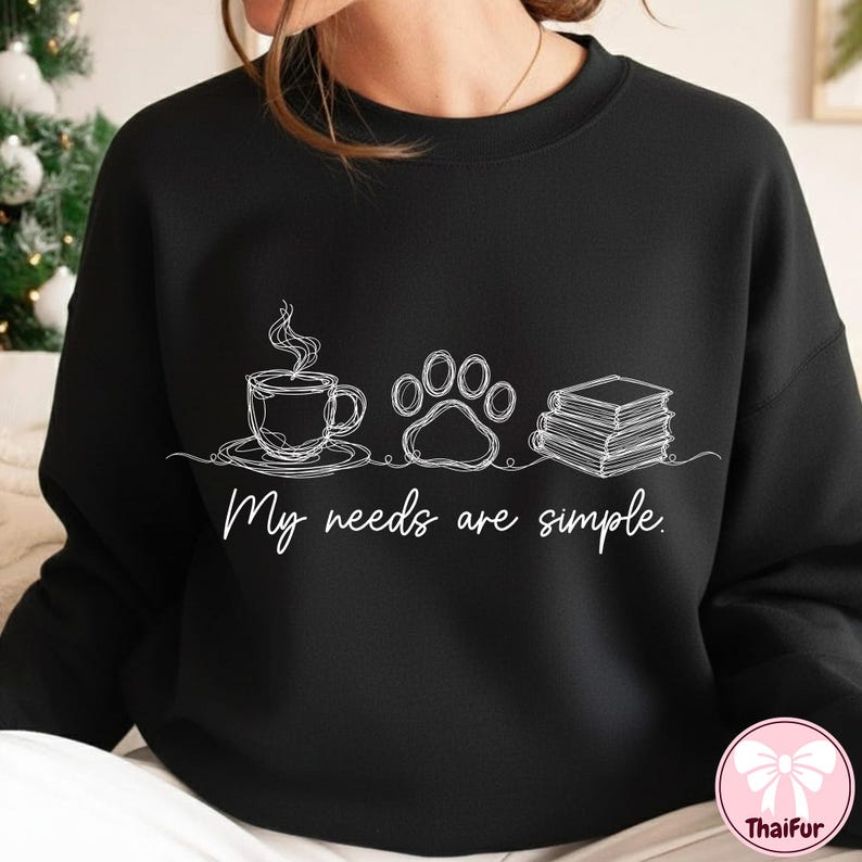 May include: Black sweatshirt with a white line drawing of a coffee cup, paw print, and books. The text "My needs are simple" is below the drawings. The sweatshirt has a crew neck.