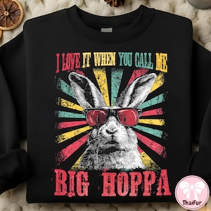 Puede incluir: Sudadera negra con un gráfico de un conejo con gafas de sol rojas. El texto "I LOVE IT WHEN YOU CALL ME BIG HOPPA" está en letras rojas y amarillas, con un diseño retro de rayos de sol.