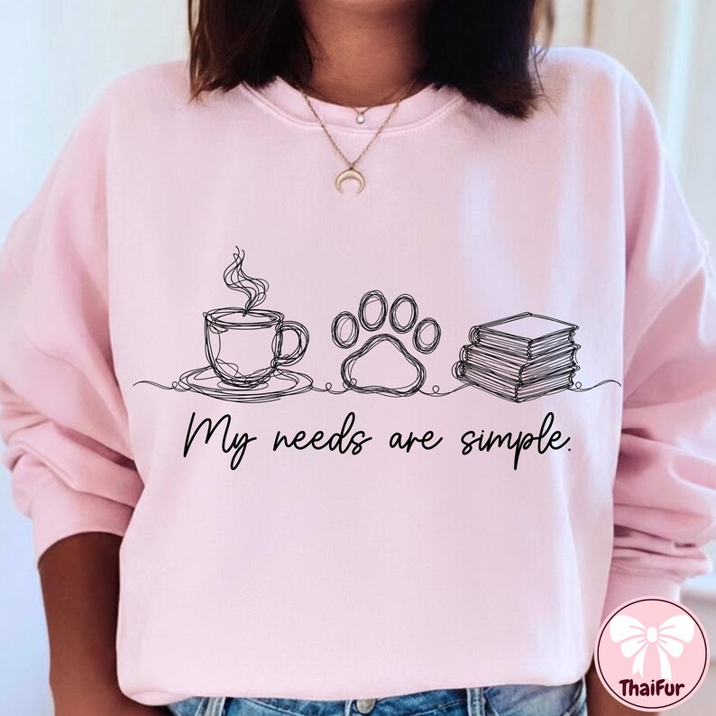 May include: A light pink sweatshirt with a minimalist line drawing of a coffee cup, paw print, and books. The text "My needs are simple" is below the drawings. The sweatshirt is a comfortable, casual style.