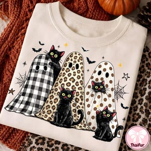 May include: A cream-colored crewneck sweatshirt featuring a graphic of three black cats wearing ghost costumes. One ghost has a black and white buffalo plaid pattern, one has a leopard print, and one has brown polka dots. Bats and spiderwebs surround them.