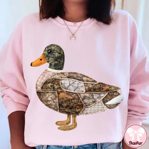 May include: A light pink sweatshirt featuring a large, appliqued duck design. The duck has a camouflage head, a brown and tan body, and an orange beak. The duck's body is made of various fabric patterns. The sweatshirt is a casual style.
