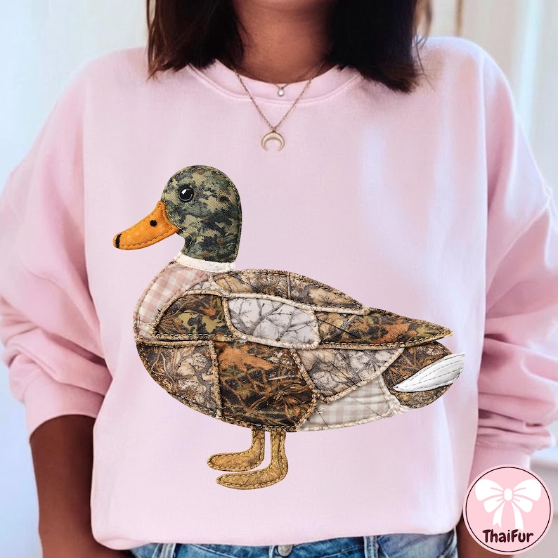 May include: A light pink sweatshirt featuring a large, appliqued duck design. The duck has a camouflage head, a brown and tan body, and an orange beak. The duck's body is made of various fabric patterns. The sweatshirt is a casual style.