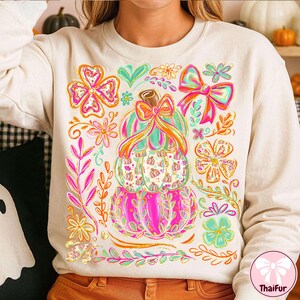 May include: Cream-colored sweatshirt featuring a stacked pumpkin design with a bow and floral accents. The pumpkins are pink, green, and white, with orange and pink bows. Surrounding the pumpkins are colorful flowers and leaves.