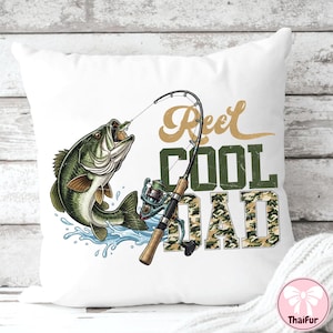 May include: White square pillow with a fishing-themed design. A green and brown fish leaps from blue water, with a fishing rod and reel. Text reads "Reel Cool Dad" in gold and camouflage.