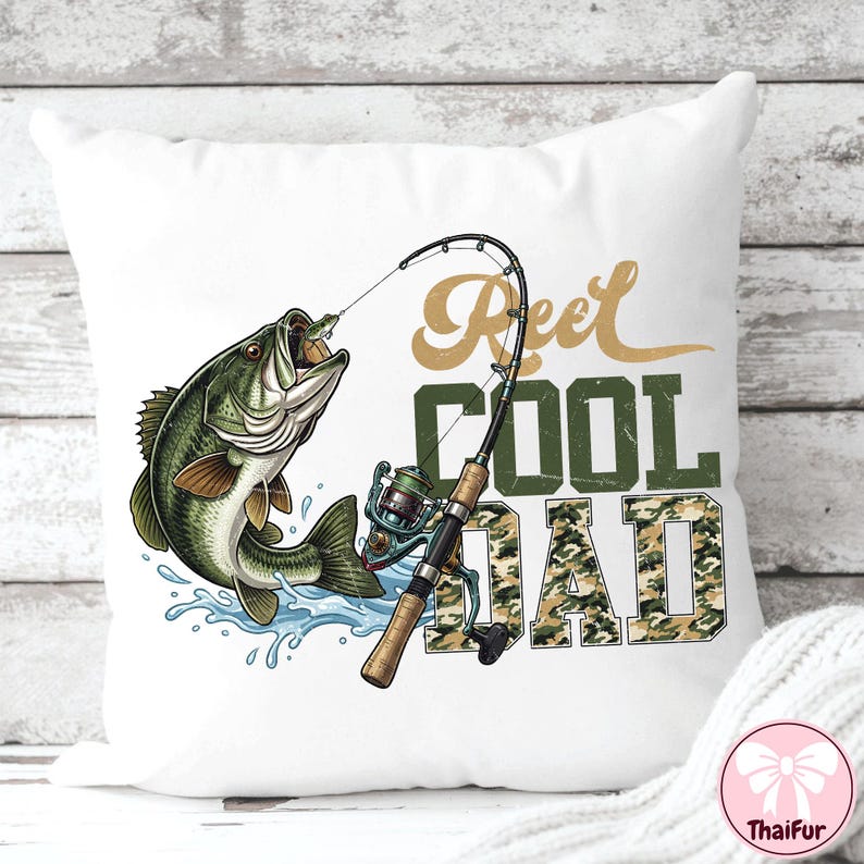 May include: White square pillow with a fishing-themed design. A green and brown fish leaps from blue water, with a fishing rod and reel. Text reads "Reel Cool Dad" in gold and camouflage.