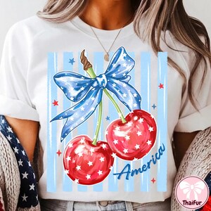 May include: White t-shirt featuring a patriotic design. The graphic includes two red cherries with white star accents, a blue bow with white stars, and the word "America" in blue script. The background has blue vertical stripes.