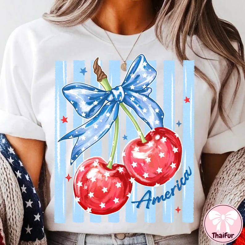 May include: White t-shirt featuring a patriotic design. The graphic includes two red cherries with white star accents, a blue bow with white stars, and the word "America" in blue script. The background has blue vertical stripes.
