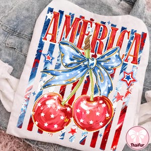 May include: White t-shirt with a patriotic design featuring the word "AMERICA" in red and gold, a blue bow, and two cherries with white stars. Red and blue stripes and stars complete the design.