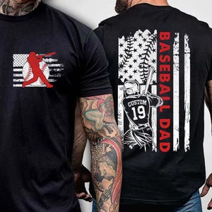 Puede incluir: Camiseta negra con un gráfico de jugador de béisbol rojo en la parte delantera y un gráfico de jugador de béisbol blanco con una bandera estadounidense y el texto "Baseball Dad" en la parte posterior.