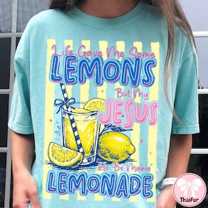 Può includere: T-shirt azzurra con grafica a tema limonata e la scritta "Life Gave Me Some Lemons But My Jesus He Be Makin' Lemonade". Il design utilizza colori giallo e blu.