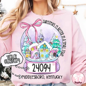 Op de afbeelding: Een lichtroze sweatshirt met een sneeuwbolontwerp. Het ontwerp bevat huizen, bomen en een sneeuwpop, met de tekst "Christmas is better in a small town." De tekst "Your Custom" is ook zichtbaar.