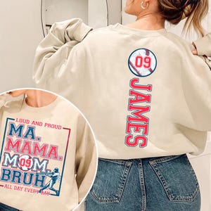 May include: A beige sweatshirt with a baseball graphic and the name "JAMES" printed vertically down the back. The front of the sweatshirt has a graphic with the text "LOUD AND PROUD MA. MAMA. MOM BRUH ALL DAY EVERY DAY" and a baseball player illustration.