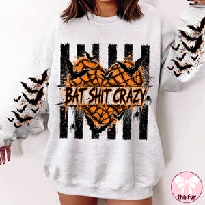 May include: A light gray crewneck sweatshirt featuring a central graphic of an orange heart filled with a spiderweb pattern, overlaid with the text BAT SHIT CRAZY. The design includes black vertical stripes, flying bats, and spiders on the sleeves.