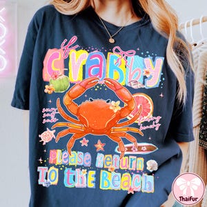 May include: Dark blue t-shirt with a vibrant graphic. The design includes an orange crab, floral accents, and the phrase "If Crabby Please Return To The Beach". The shirt has a loose fit and a summery theme.