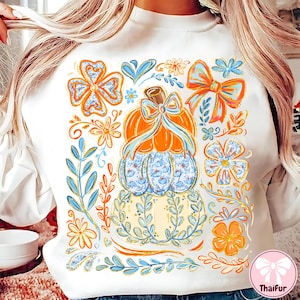 May include: White sweatshirt featuring a colorful fall-themed design. The design includes an orange pumpkin with a blue bow, surrounded by flowers, leaves, and bows in shades of orange, blue, and yellow. The sweatshirt has a crew neck.