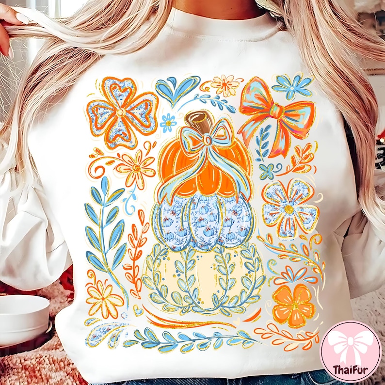 May include: White sweatshirt featuring a colorful fall-themed design. The design includes an orange pumpkin with a blue bow, surrounded by flowers, leaves, and bows in shades of orange, blue, and yellow. The sweatshirt has a crew neck.