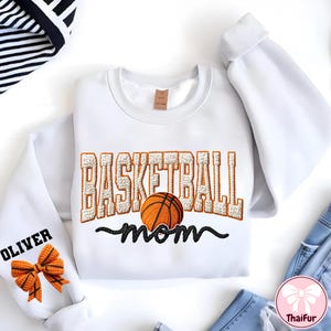 Puede incluir: Sudadera blanca con el texto bordado "BASKETBALL mom" y un gr&aacute;fico de baloncesto. La palabra "OLIVER" est&aacute; bordada en la manga izquierda, junto con un lazo en forma de baloncesto. La sudadera tiene mangas largas.