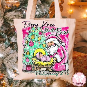 May include: A light pink tote bag featuring a Christmas scene with Santa Claus, a baby, and a Christmas tree. The text "Every Knee Shall Bow" and "Philippians 2:10" are also displayed on the bag.