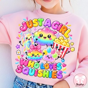 May include: A light pink sweatshirt featuring a colorful graphic print with the text JUST A GIRL WHO LOVES SQUISHIES. The design includes cartoon characters of a butter stick, a rainbow cloud, a bow-topped squishy, a paw, and a popcorn bucket.
