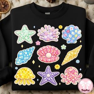 May include: Black sweatshirt featuring colorful seashell and starfish designs. The designs include a green starfish, a purple hand-shaped shell, and a blue seashell, all with floral patterns. Additional small flowers and diamonds are scattered around the designs.